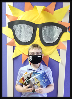 Student in a mask holding several books, standing in front of a picture of the sun. 